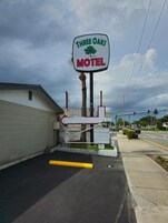 Letrero de motel con logotipo de árbol, una flecha direccional y un estacionamiento con bordillo amarillo.