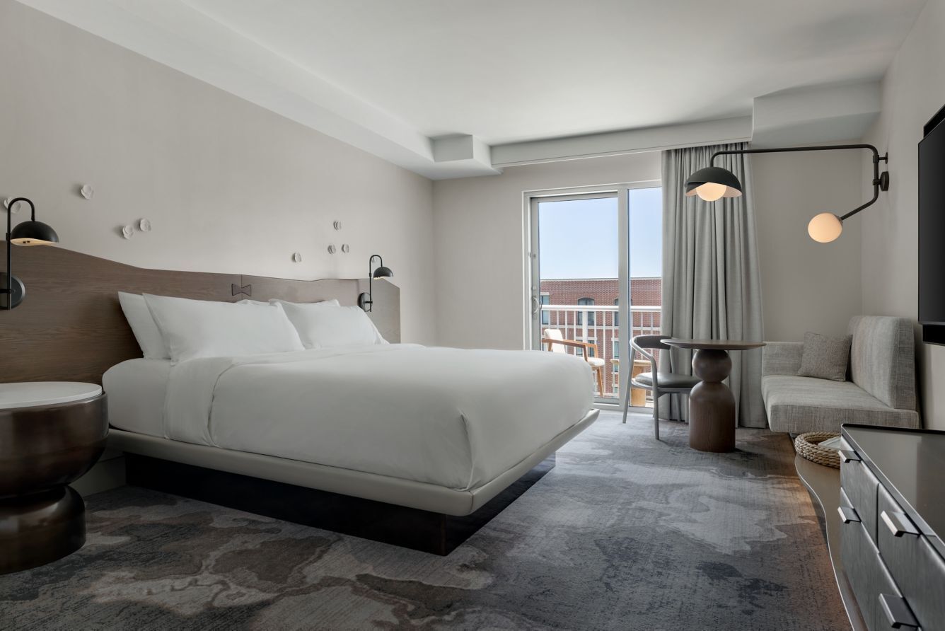 A modern hotel room with a large bed, bedside tables, a dresser, a balcony with a view, and a ceiling light fixture.