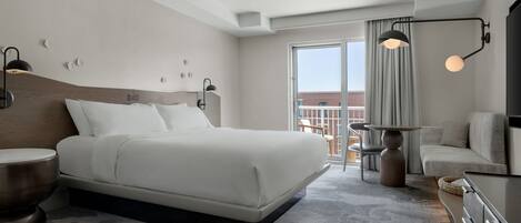 A modern hotel room with a large bed, bedside tables, a dresser, a balcony with a view, and a ceiling light fixture.