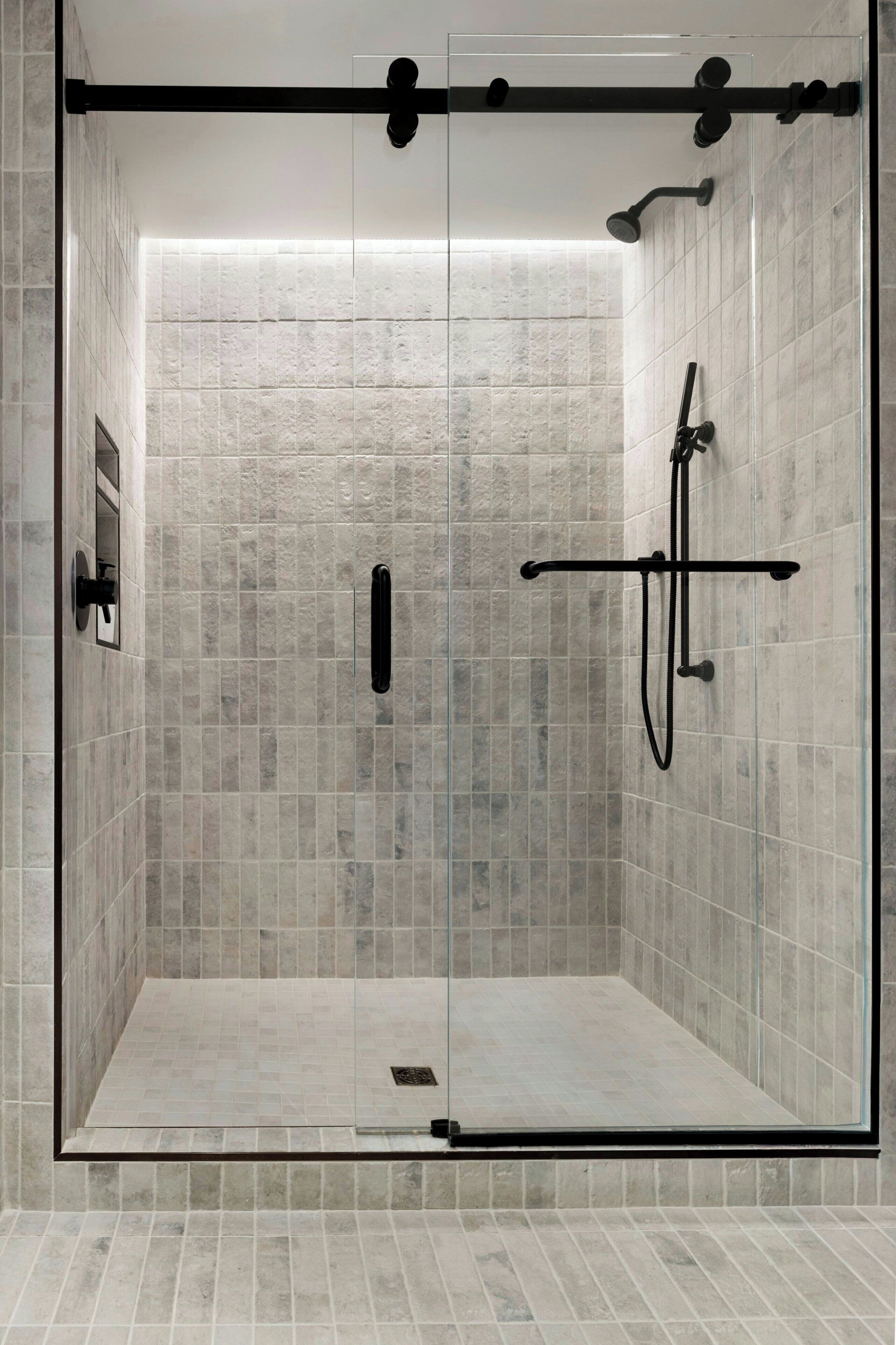 A glass-enclosed shower with a tiled floor and walls, a handheld showerhead, and a handheld faucet.