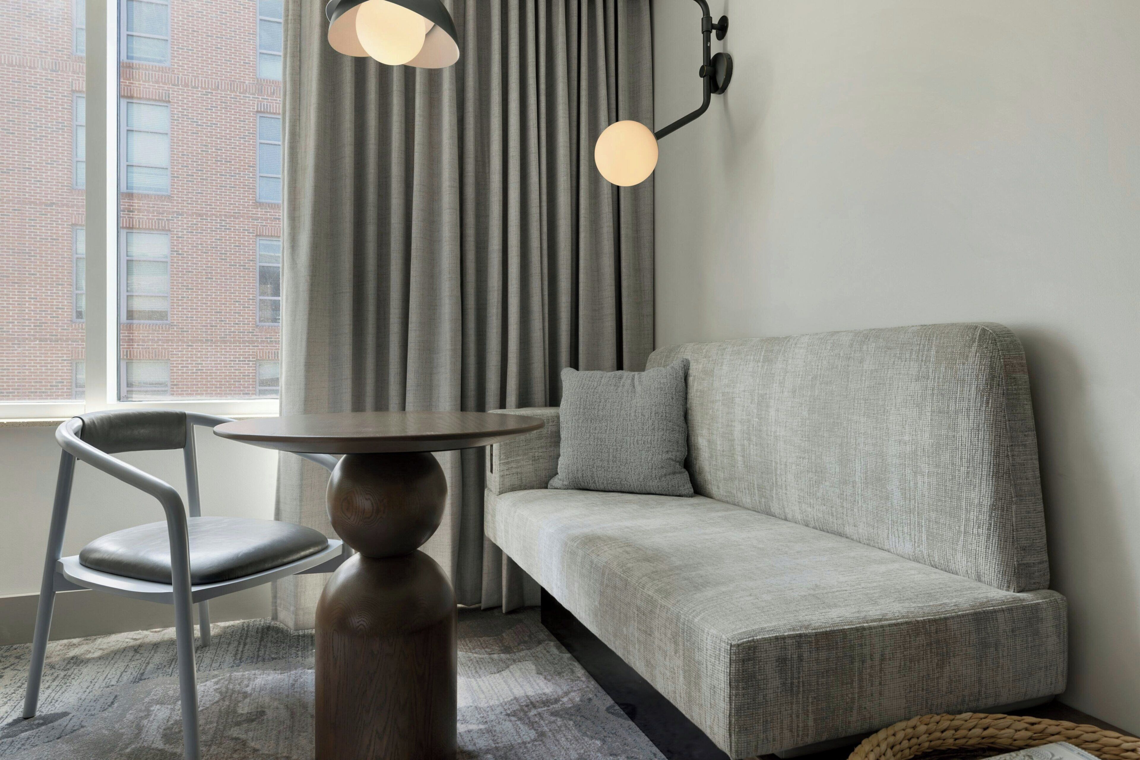 Une pièce moderne avec un canapé gris, une table ronde en bois, une chaise grise et une fenêtre avec des rideaux.