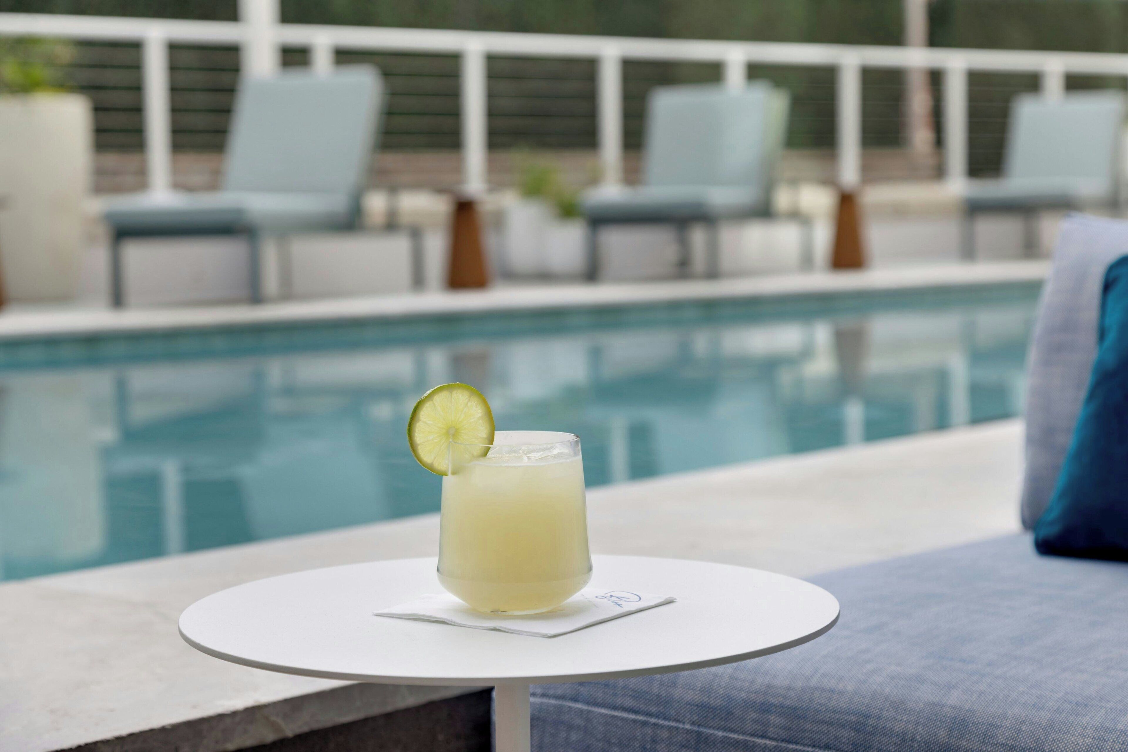 A glass of drink with a lime garnish on a white table by a poolside lounge area.