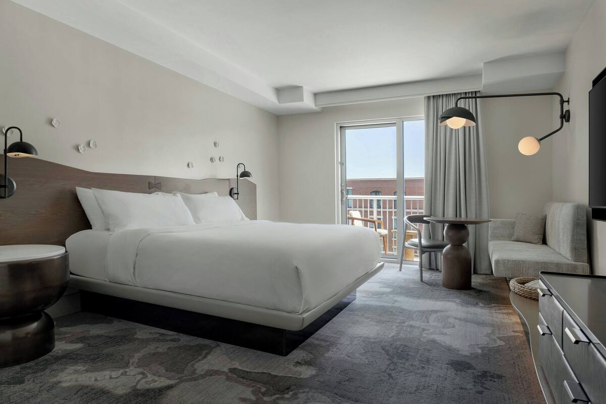 A modern hotel room with a large bed, a desk, a chair, a TV, and a balcony with a view.