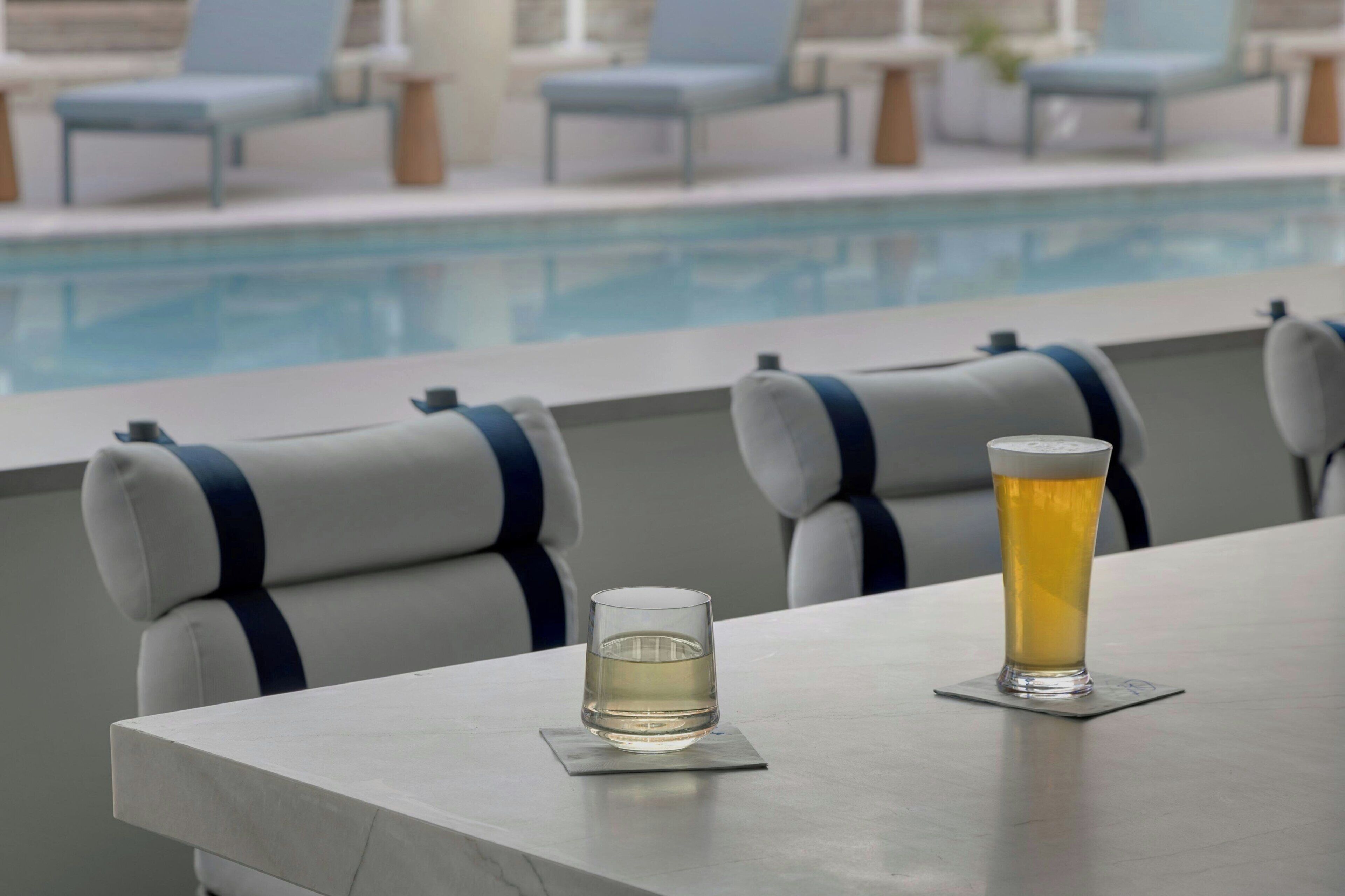A poolside bar with two glasses of beer, a pool, and lounge chairs in the background.