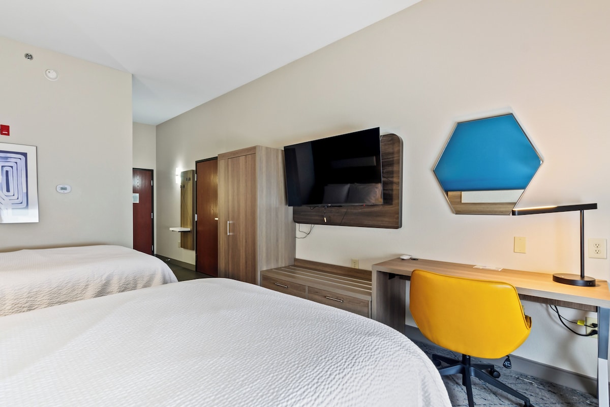 Quarto de hotel com duas camas, uma secretária com cadeira, televisão na parede e um guarda-roupa.