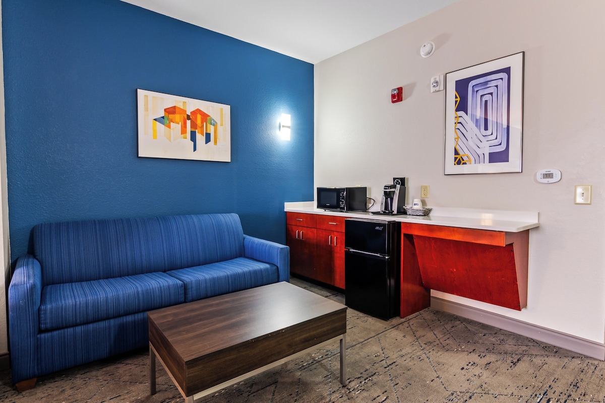 Um espaço de estar compacto com um sofá azul, uma mesa de centro de madeira, uma kitchenette com micro-ondas e frigorífico, e duas obras de arte suspensas na parede.