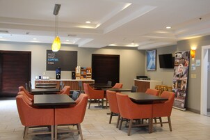 A modern cafe with orange chairs and tables, a coffee station, and a flat-screen TV on the wall.