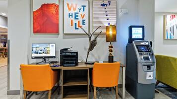 Un hall de hotel con mostrador de recepción, dos sillones naranja, un monitor de computadora, un cajero automático y obras de arte en las paredes.