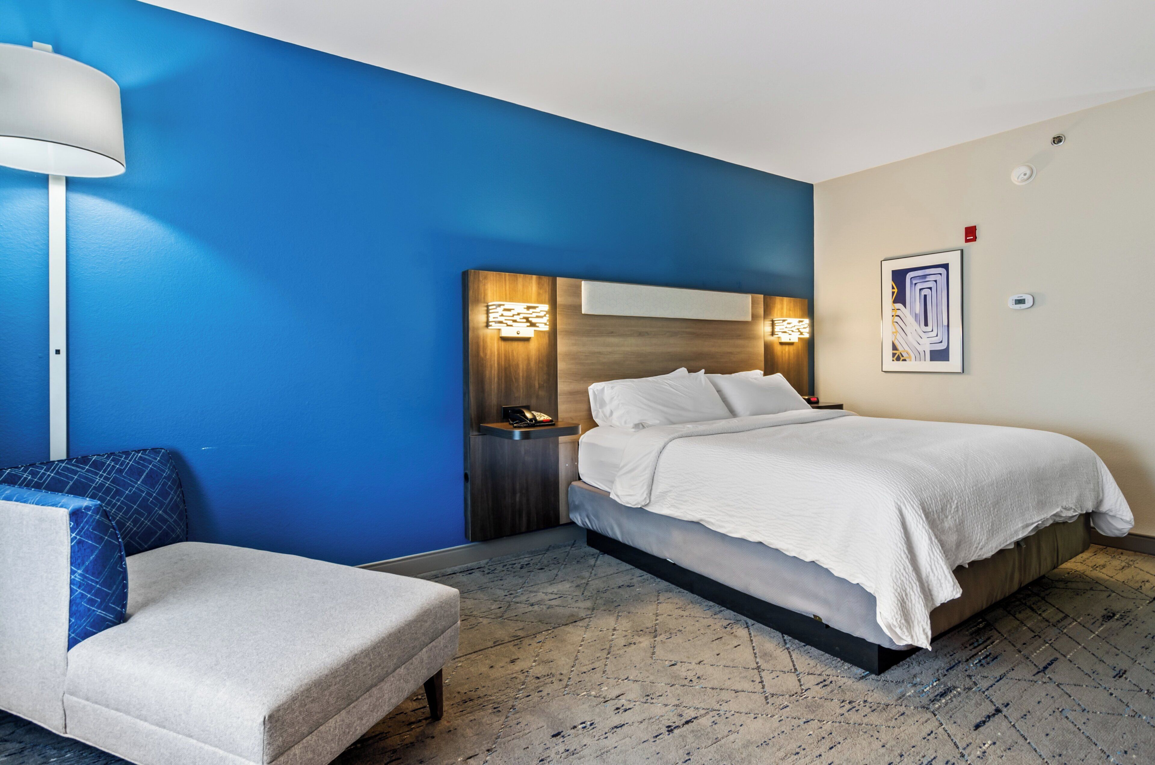 A modern hotel room with a blue accent wall, a large bed with white linens, a bedside table with a lamp, and a small chair.