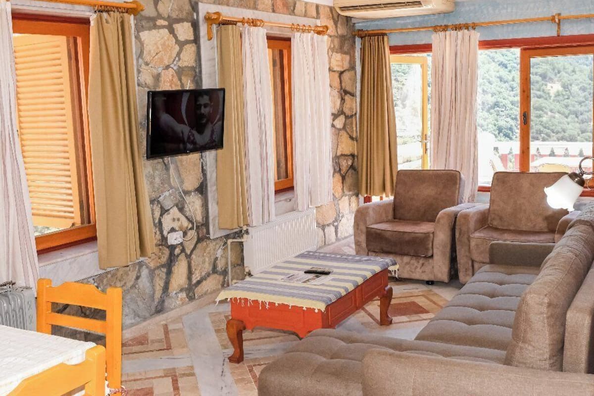 A living room with stone walls, a sofa, armchairs, a TV, and a table with chairs.