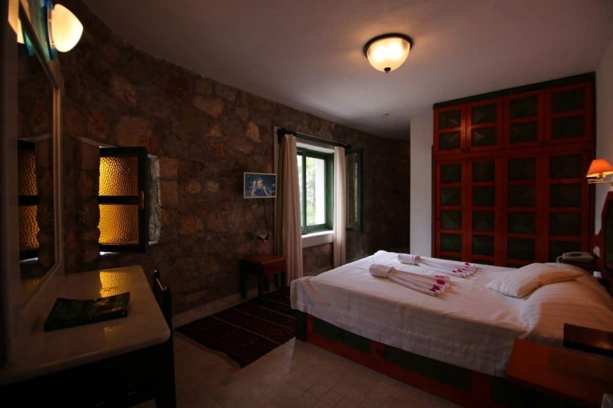 A bedroom with a stone wall, a bed with towels, a desk, and a lamp.