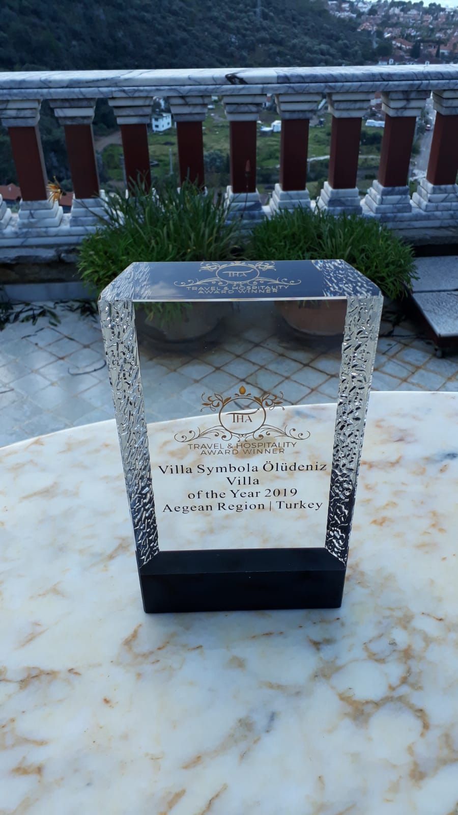 A clear award plaque on a marble table with a view of a balcony and greenery.