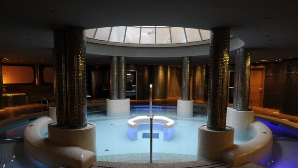 A modern indoor pool with a central circular seating area, surrounded by tall pillars, and a skylight above. - Cumbria Spa&hotel (Ciudad Real)