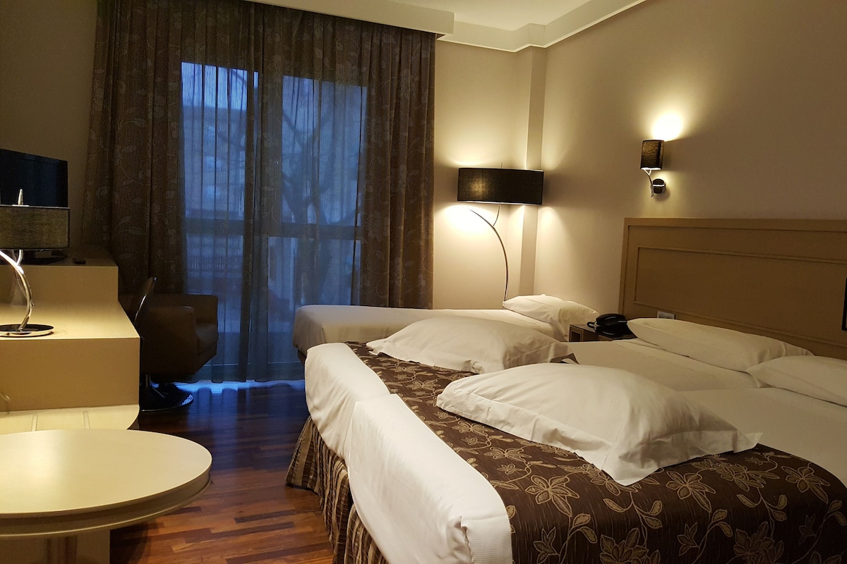 Ein Hotelzimmer mit zwei Betten, einem Schreibtisch, einem Stuhl und einem großen Fenster mit Vorhängen.
