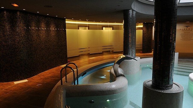 A modern indoor pool area with a curved design, a glass wall, and a tiled floor. - Cumbria Spa&hotel (Ciudad Real)
