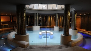 A modern indoor pool with a central circular seating area, surrounded by tall pillars, and a skylight above. - Cumbria Spa&hotel (Ciudad Real)