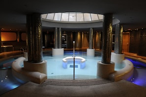 Piscina interior moderna com área de estar circular central, rodeada por pilares altos, e uma clarabóia acima.