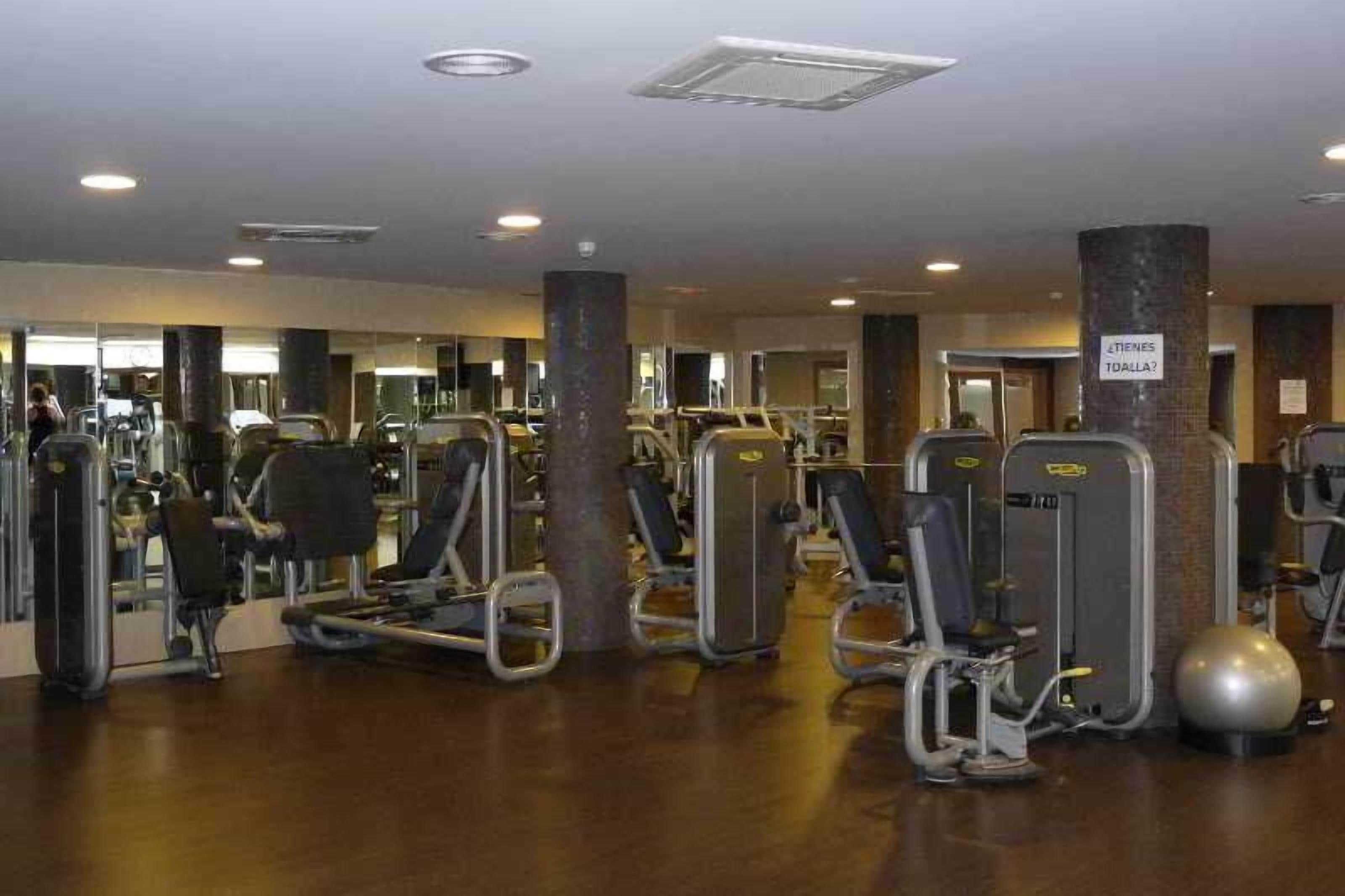 Un gimnasio con diversas máquinas de ejercicio, un espejo grande y un letrero que dice 'Tienes toalla?'.