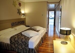Habitación de hotel con una cama grande, una mesita de noche con lámpara, y un pasillo que conduce a una puerta.