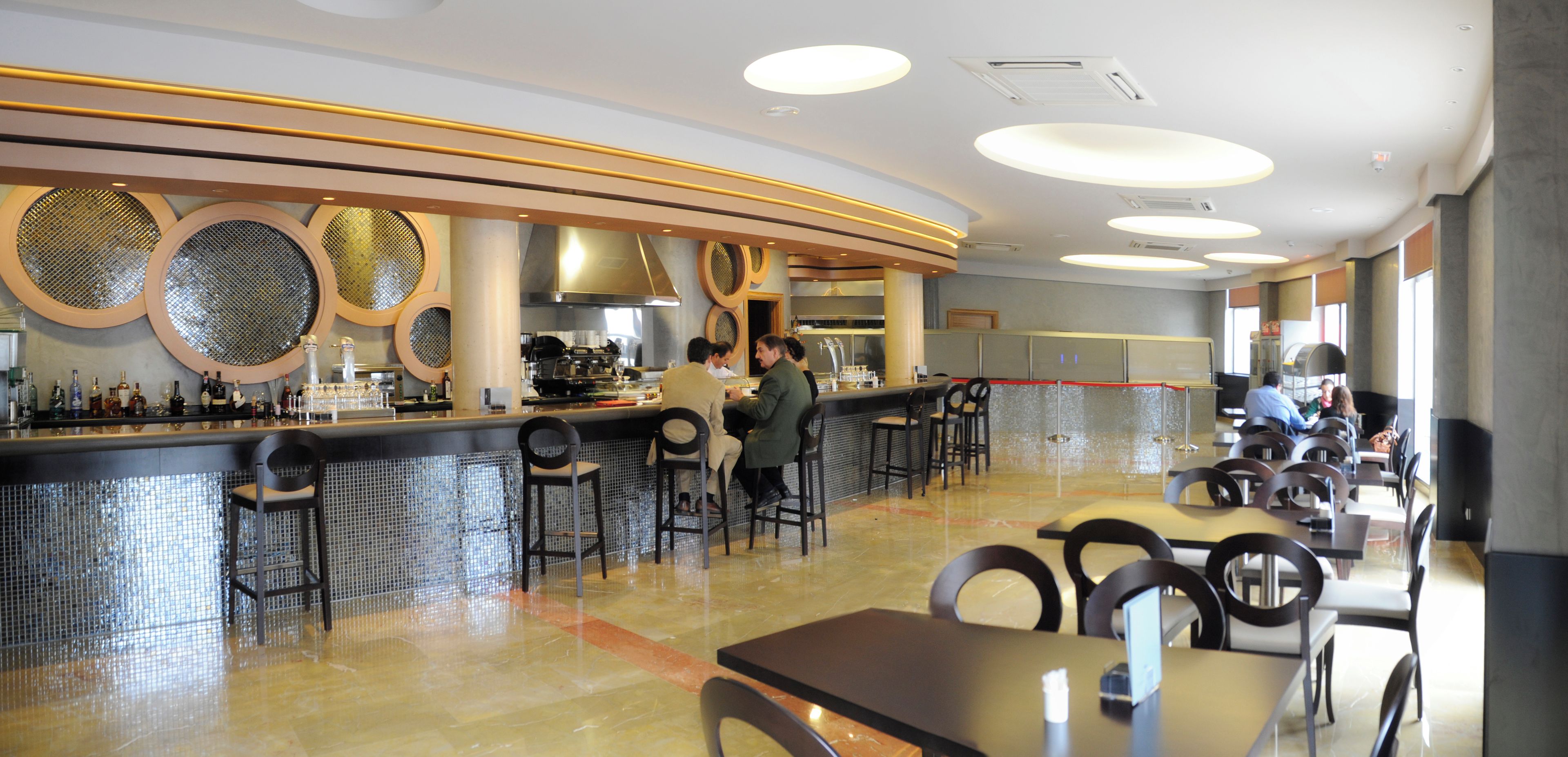 Ein modernes Restaurant mit Barbereich, Sitzgelegenheiten und einer sichtbaren Küche im Hintergrund.
