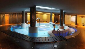 A large indoor pool with multiple hot tubs and lounge areas, surrounded by stone walls and columns. - Cumbria Spa&hotel (Ciudad Real)