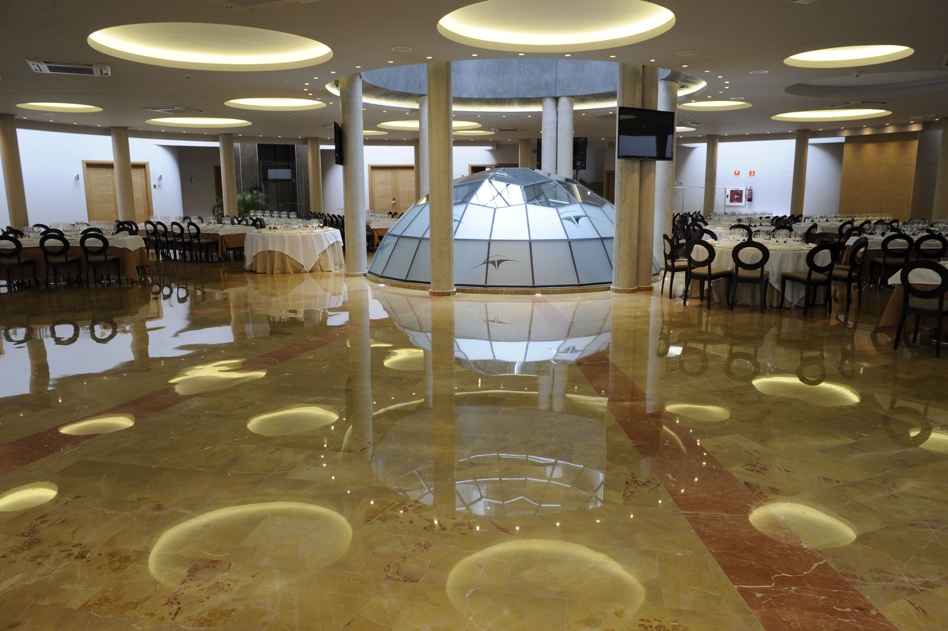 Un amplio salón con una estructura central de cúpula de cristal, varias mesas con sillas y un suelo de mármol pulido.
