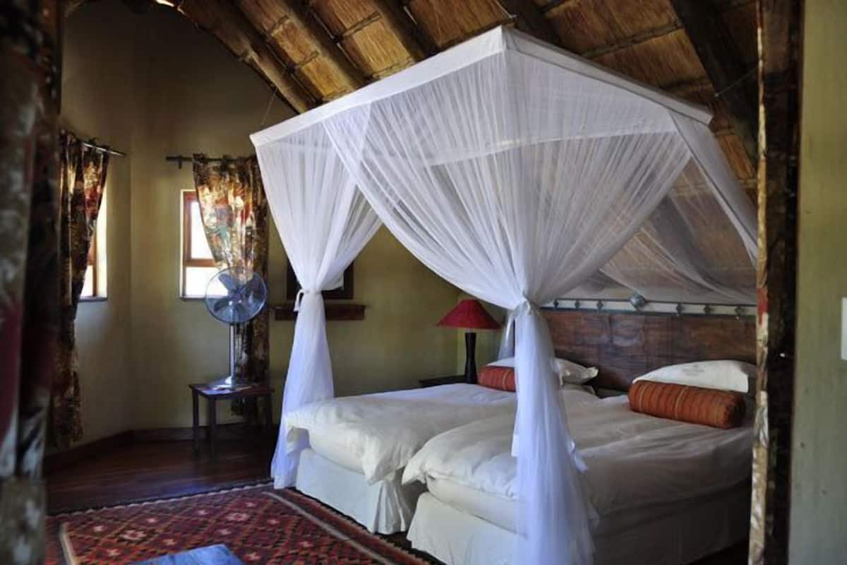 A room with two beds under a thatched roof, a fan, and a patterned rug.