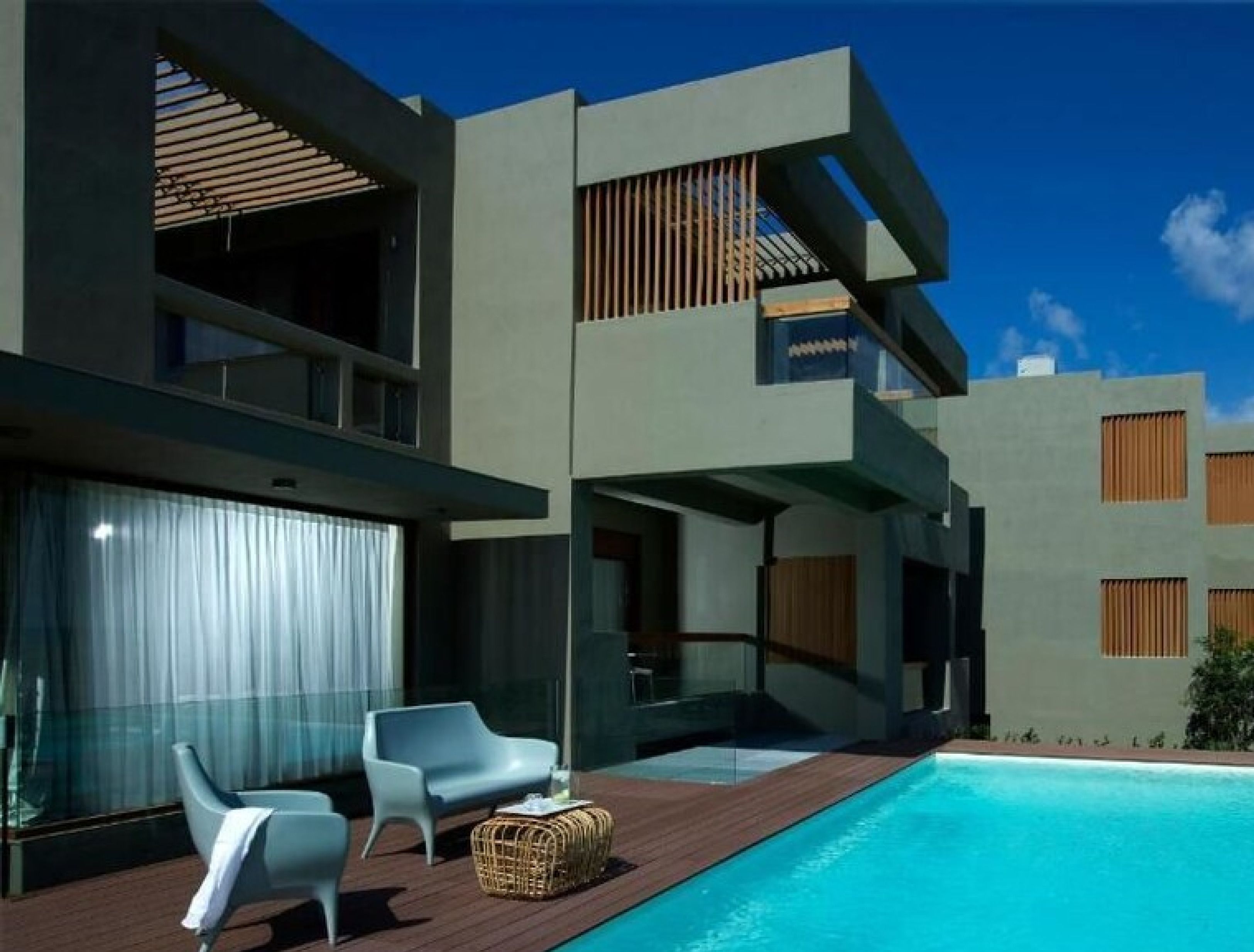 Un bâtiment moderne doté d’une piscine, caractérisé par une façade en verre, des chaises blanches et une terrasse en bois.
