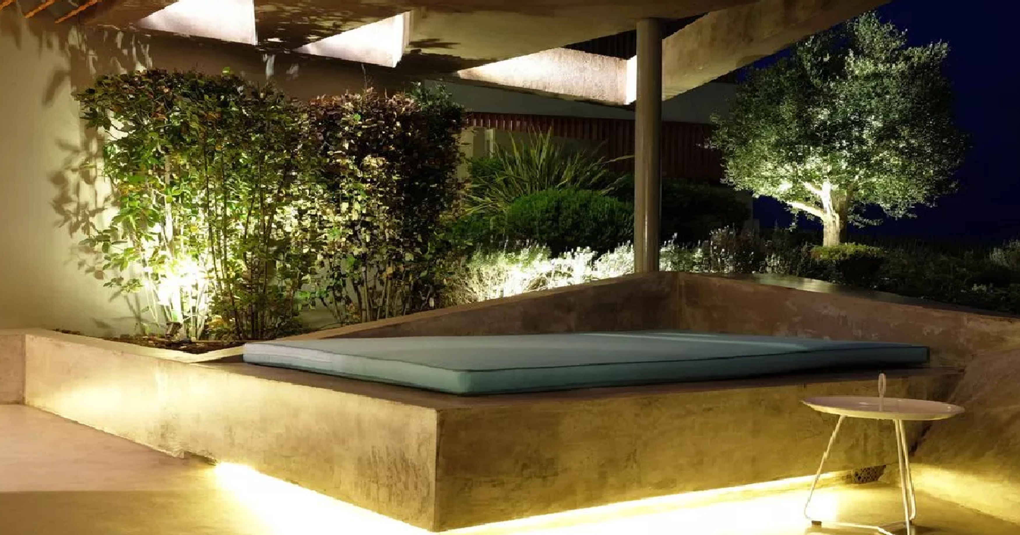 An outdoor seating area with a cushioned bench and a small table, surrounded by lush greenery and a tree.