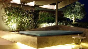 An outdoor seating area with a cushioned bench and a small table, surrounded by lush greenery and a tree.