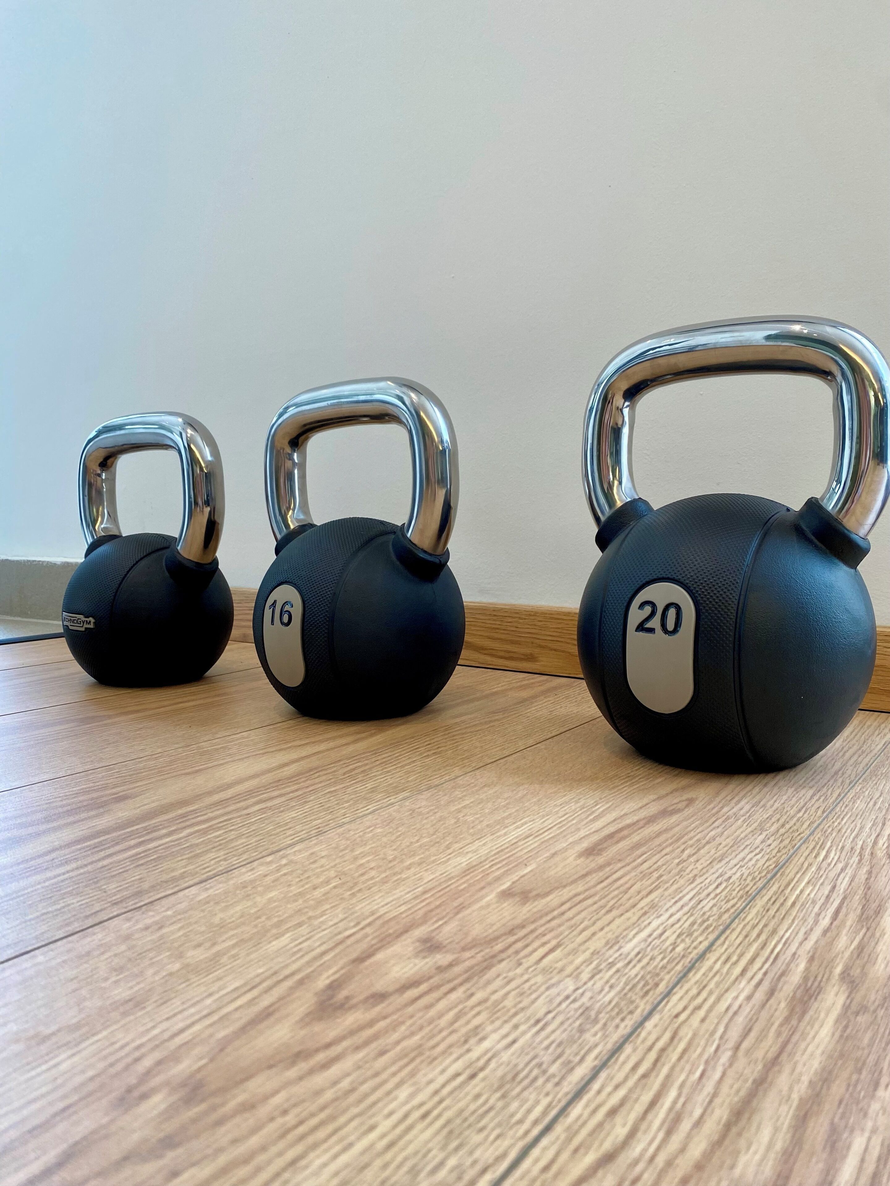 Trois kettlebells, dont les poids sont de 16 kg et 20 kg, et un troisième dont le poids n’est pas précisé, sont posés sur un plancher en bois.