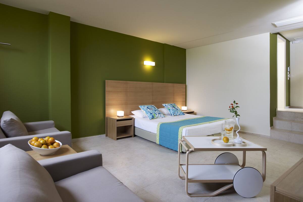 A modern hotel room with a bed, a bedside table with a lamp, a small table with a drink tray, a bowl of fruit, and a sofa.