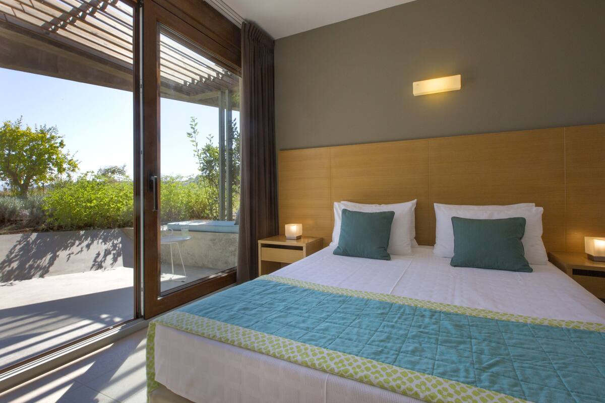 A large bed with a quilt, two bedside tables, a nightstand, and a window with a view of greenery.