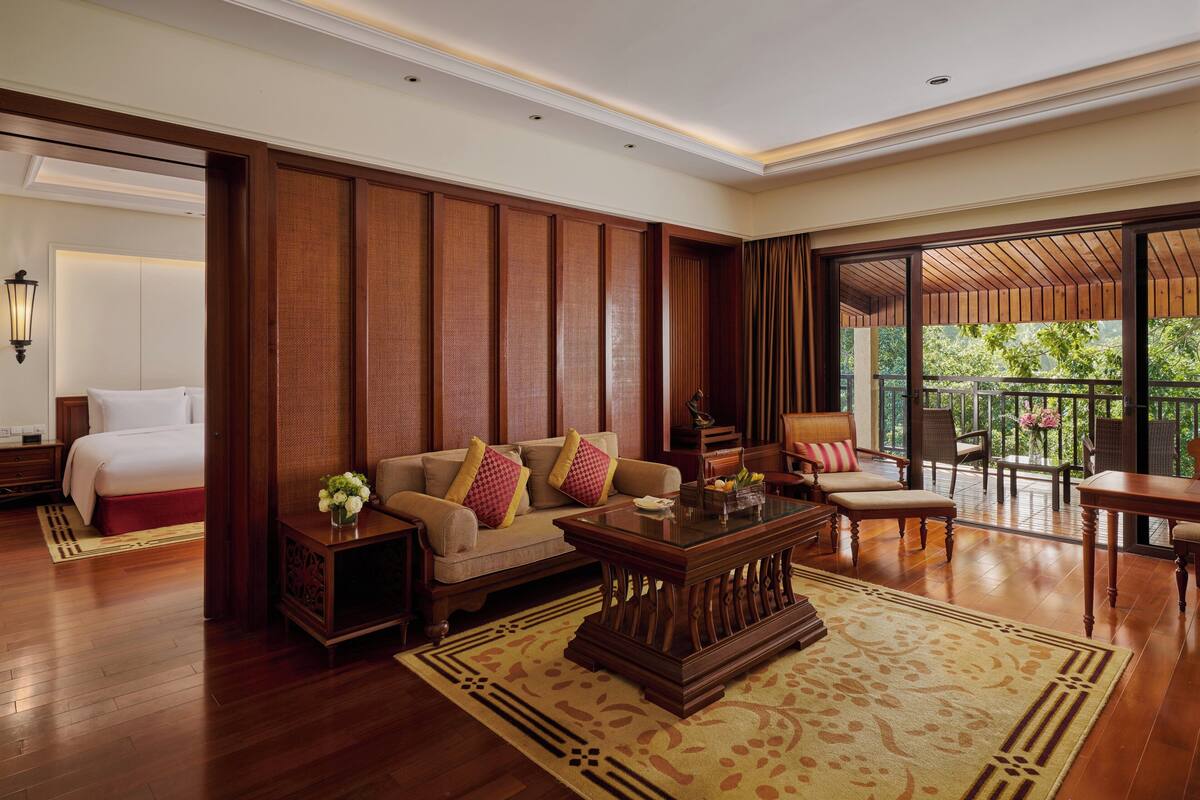 A spacious living area with a sofa, coffee table, and chairs, a balcony with a view of greenery, and a neatly arranged bedroom with a bed and nightstand.