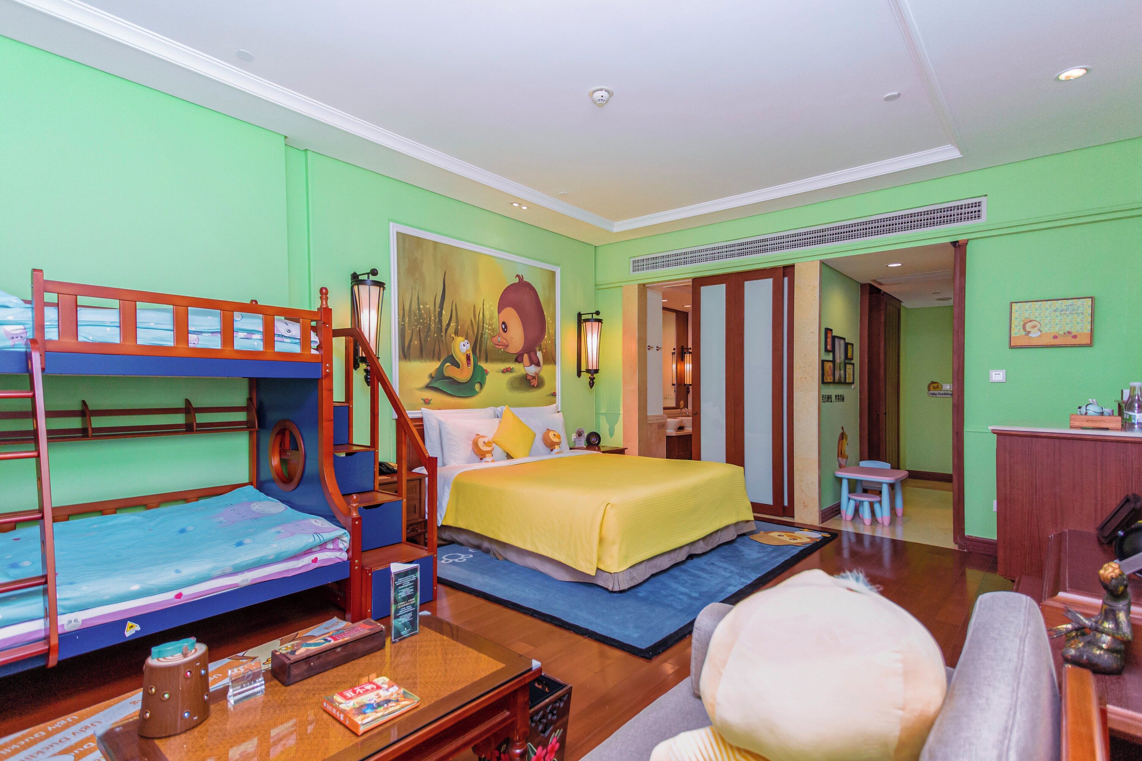 A hotel room with bunk beds, a bed with a yellow blanket, a desk with a lamp, and a small table with books.