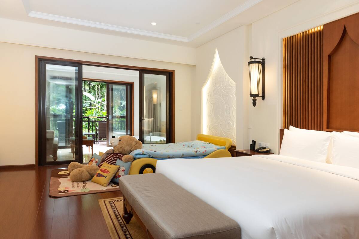 A hotel room with a large bed, a smaller bed, a sofa, a teddy bear, and a sliding glass door leading to an outdoor area.