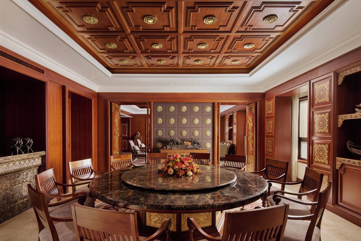 A formal dining room with a round marble table, wooden chairs, and a decorative ceiling.