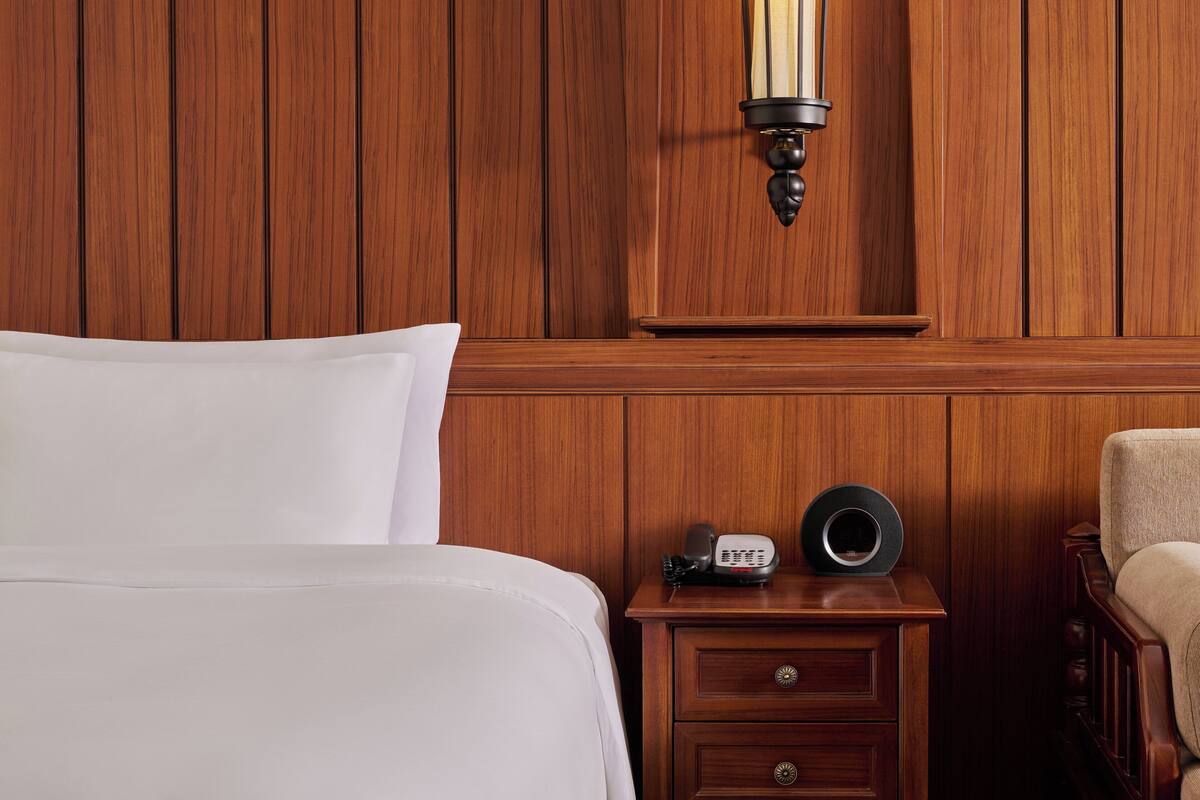 A wooden-paneled room with a bed, a nightstand with a telephone and a speaker, and a chair.