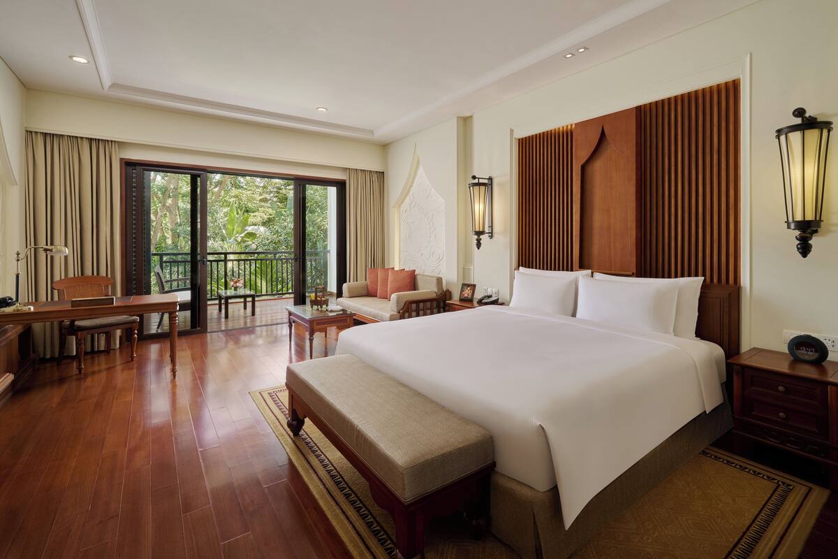 A spacious hotel room with a large bed, a balcony with a view of greenery, a dining area with a table and chairs, and a small seating area with a sofa and a lamp.