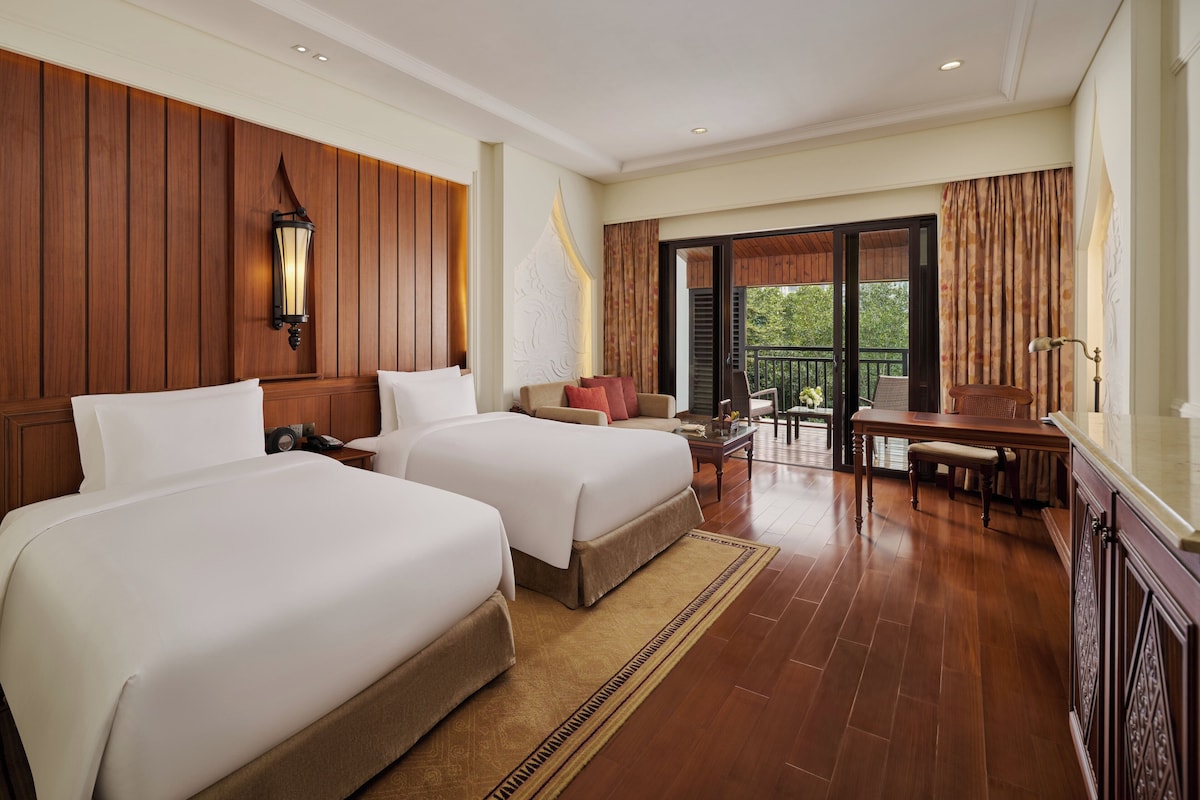 A hotel room with two beds, a sitting area with a table and chairs, a balcony with curtains, and a wooden wall panel.