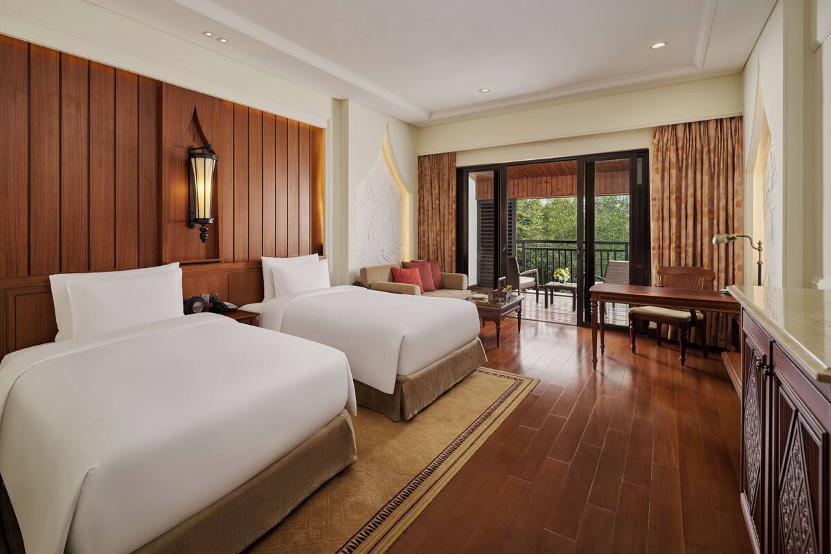 A hotel room with two beds, a sitting area with a table and chairs, a balcony with curtains, and a wooden wall panel.