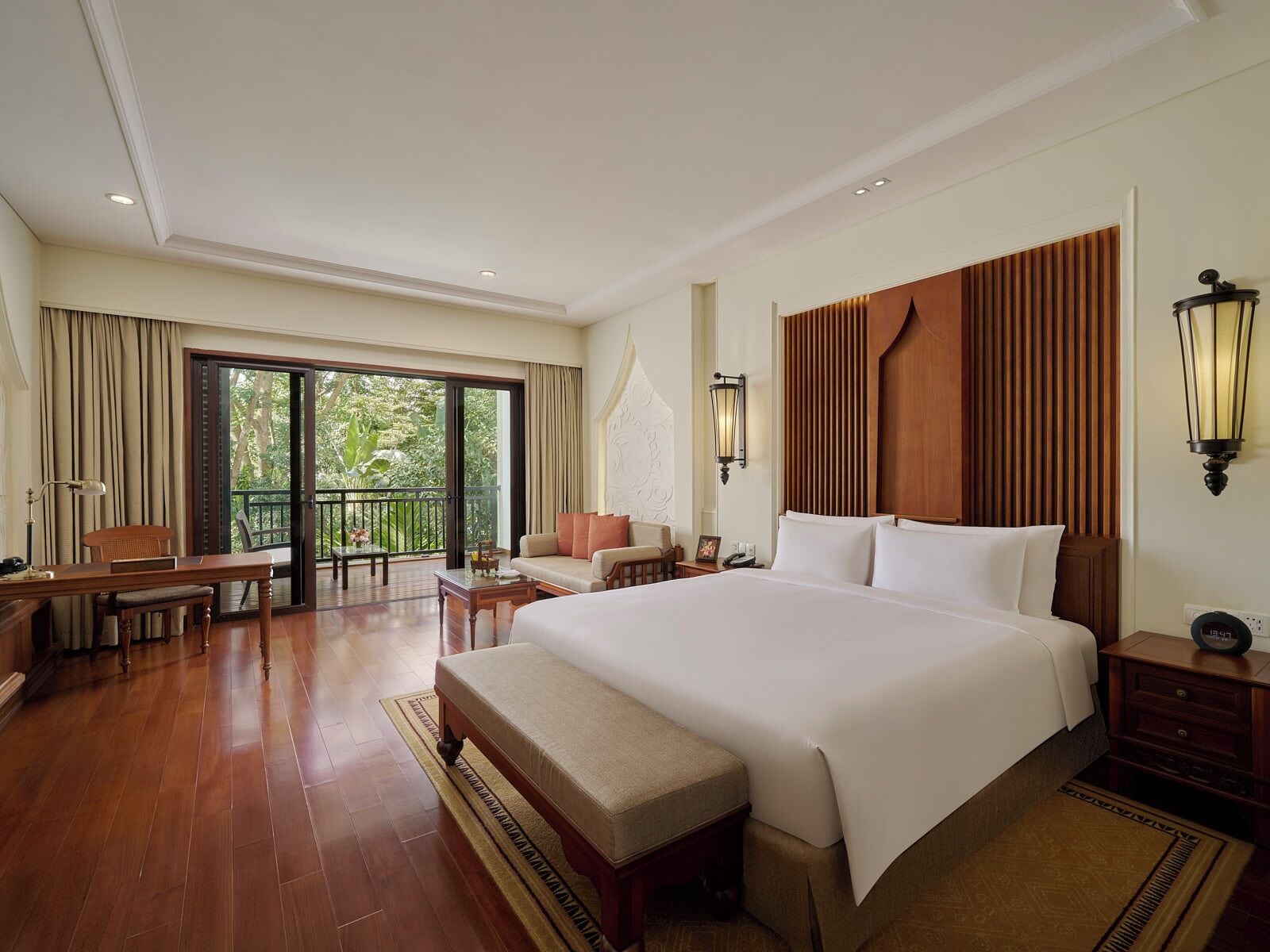 A hotel room with a large bed, a desk, a chair, a bench, a nightstand, a lamp, and a view of the outdoors through a sliding door.