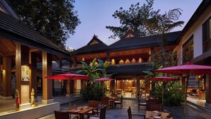 Un espacio de comedor al aire libre con mesas y sillas de madera, sombrillas rojas y un estilo arquitectónico tradicional.