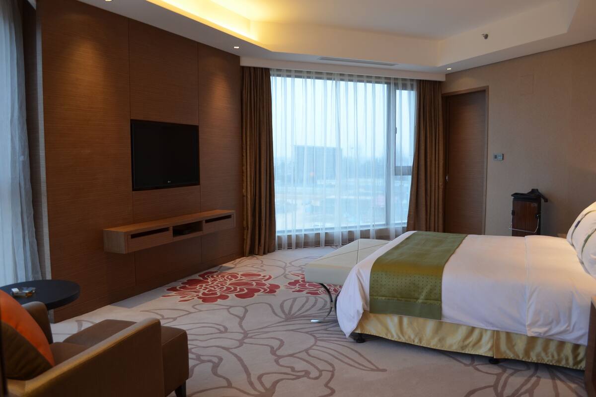 A hotel room with a bed, a TV mounted on the wall, a window with curtains, and a small table.