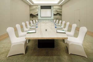 Un salón de conferencias con una larga mesa, sillas blancas y una pantalla de proyector.