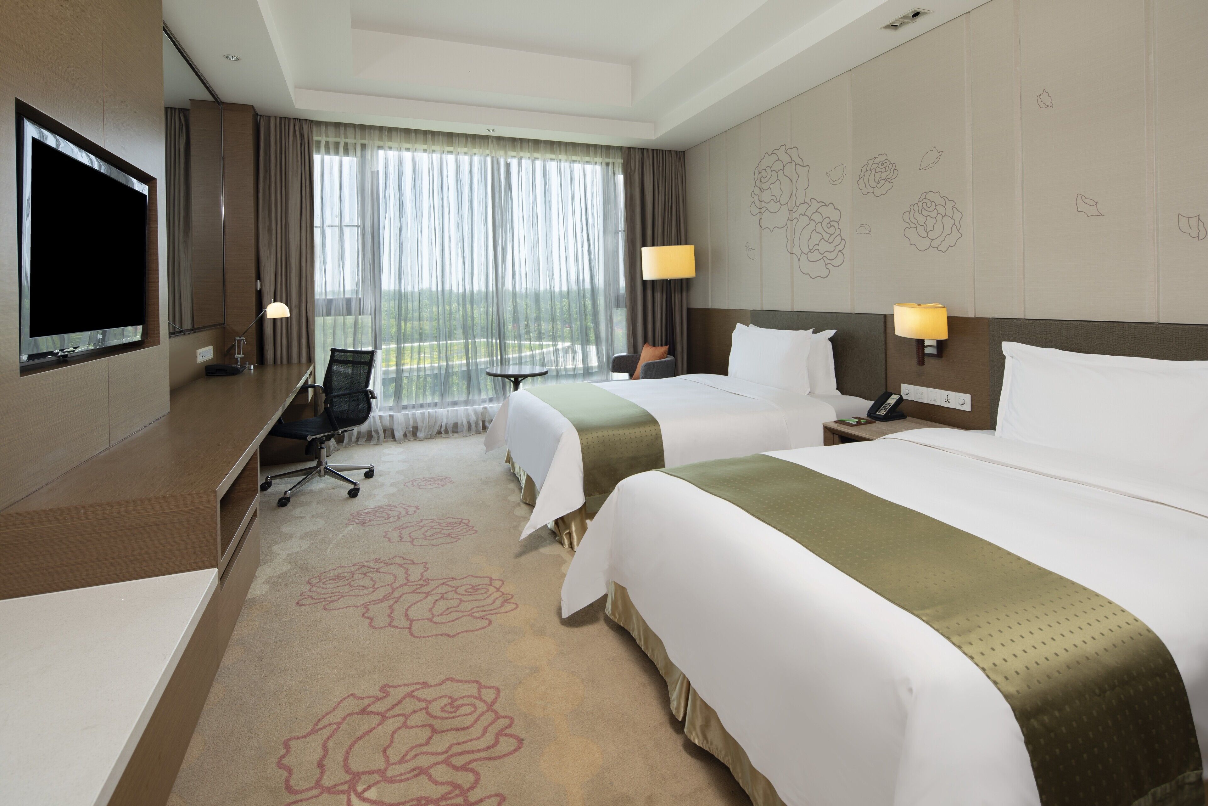 A hotel room with two beds, a desk with a chair, a TV, and a large window with curtains.