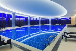 Una amplia piscina interior con diseño de mosaico azul, rodeada de sillones de descanso y un muro de cristal.