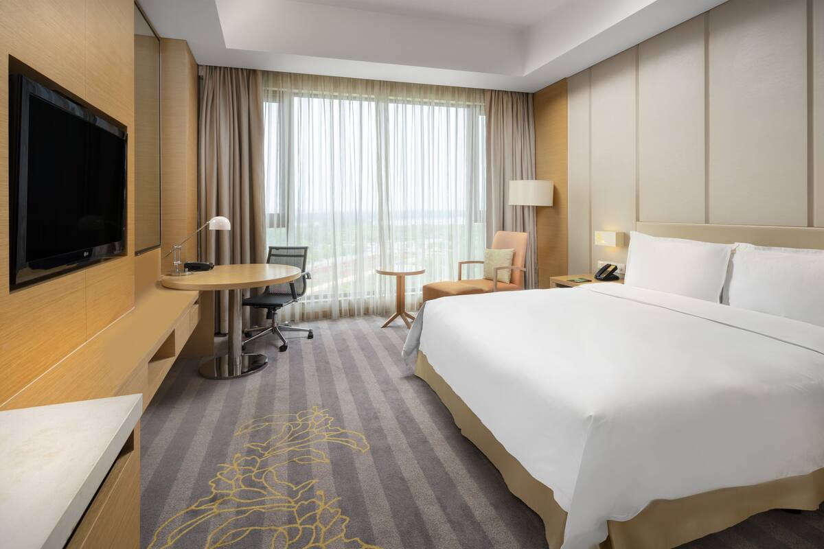Quarto de hotel com uma cama grande, uma escrivaninha com cadeira, televisão e janela com cortinas.