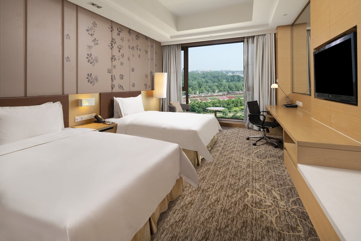 Quarto de hotel com duas camas, uma mesa, uma televisão e vista para um parque.