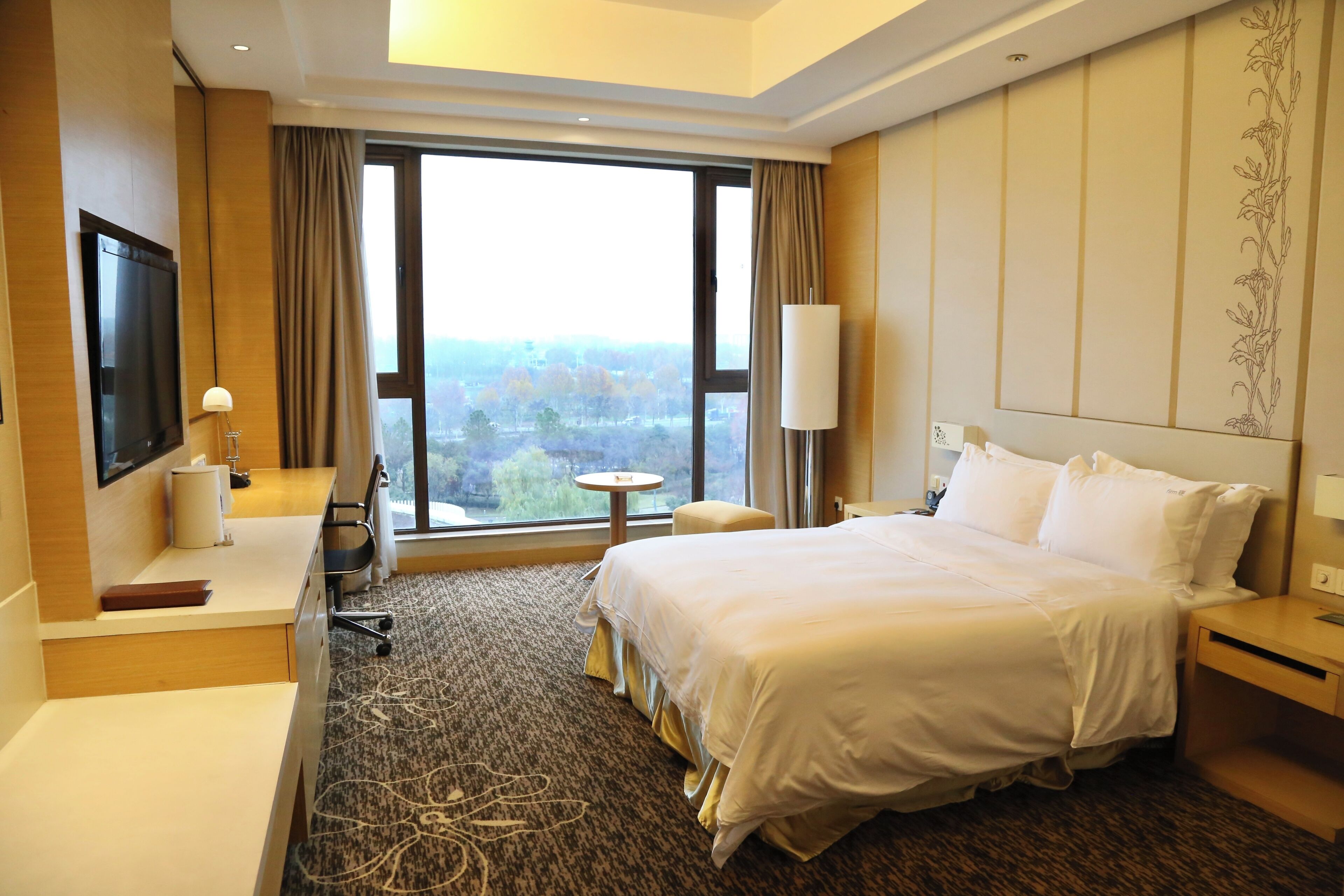 A hotel room with a large bed, a desk with a chair, a TV, and a window with a city view.